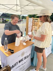A vendor hands a bag to a customer at an outdoor booth labeled "Daisy Mae Designs." The booth displays various items, including maps and a bottle of water.
