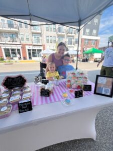 A woman and two children stand behind a market stall selling baked goods under a tent, with buildings and other market stalls in the background.