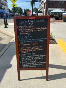A sidewalk chalkboard lists five signature drinks with ingredients, including fresh lemonade, pineapple, strawberry, mango, coconut, and apple, at an outdoor market.