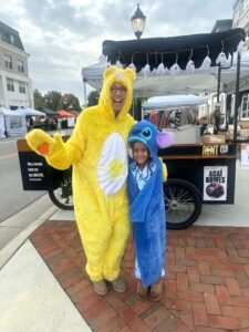 An adult in a yellow bear costume and a child in a blue Stitch costume stand together smiling at an outdoor market.