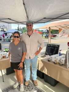 Two people stand under a market tent beside a table displaying tea products and a chalkboard menu at an outdoor market.