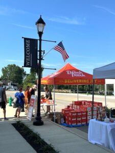 A street market scene with people standing by a red tent labeled "The Peach Truck," surrounded by boxes of peaches. An American flag and a "Welcome" sign are visible.