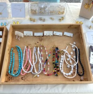 A wooden tray displays colorful beaded necklaces and pearl necklaces, organized in rows. Behind the tray are small gold earrings and jewelry cards on a white tablecloth.