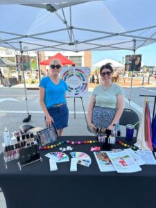 Two women stand behind a table display with cosmetics, color palettes, and brochures at an outdoor market under a canopy tent.