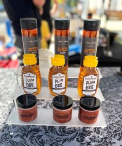 Display of Buzzin' Hot Honey bottles, Plum Prairie Plum Bee honey containers, and jars of Strawberry Lavender preserves on a clear acrylic stand over a patterned tablecloth.