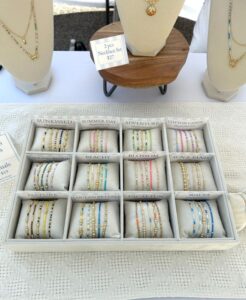 A display tray with twelve sections shows sets of colorful beaded bracelets labeled with names like "Sunkissed," "Adventure," and "Cotton Candy." Necklace displays stand behind the tray.
