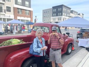 An older woman and man stand by a red vintage truck at an outdoor market; the man is eating corn while the truck bed is filled with ears of corn.