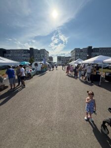Outdoor market with vendor tents and people browsing on a sunny day; a young girl stands in the foreground on a paved street lined with apartment buildings.