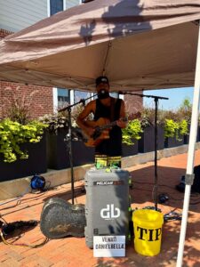 A musician plays guitar and sings under a canopy on a brick sidewalk, with a tip bucket and equipment in front of him.