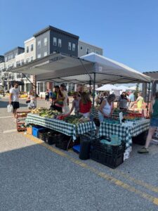 People browse and purchase fresh produce from a vendor stall at an outdoor farmers market on a sunny day.