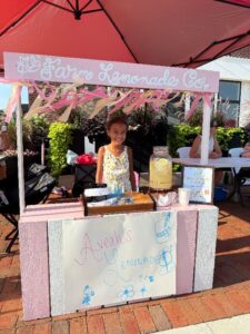 A young girl stands behind a decorated lemonade stand labeled "Avaeh's Lemonade Co." with a pitcher of lemonade, cups, and a sign, outdoors on a sunny day.