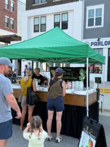 People stand in line at an outdoor market stall with a green canopy, ordering from staff at a booth advertising pickle flavors. Nearby buildings and a sidewalk are visible in the background.