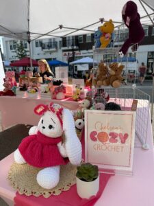 Handmade crochet stuffed animals displayed on pink tables at an outdoor market stall, with a sign reading "Chelsea's Cozy Crochet" in the foreground.