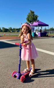 Young girl in a pink dress and headband stands on a purple and pink scooter in a parking lot, with a purple tent and trees in the background.