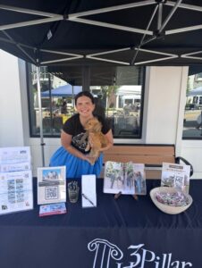 A woman stands behind an information booth for 3 Pillar with brochures, magazines, a QR code, and a bowl of candy, holding a small brown dog under a canopy.