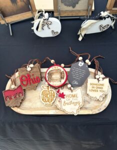 A wooden tray displays various Ohio-themed ornaments with messages like "Ohio," "Best Teacher Ever 2023," "Home of the Buckeyes 2023," and "Someone in Ohio Loves You.