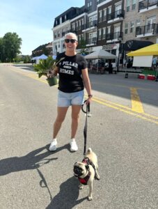 A woman in sunglasses holds a potted plant and a pug on a leash while standing on a street with shops and market tents in the background.