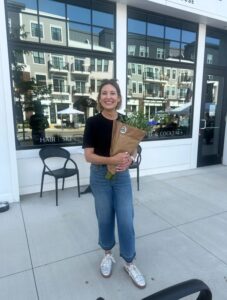 A woman stands on a sidewalk outside a building, smiling and holding a bouquet of flowers wrapped in brown paper.