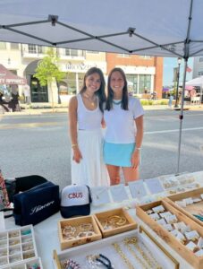Two women stand behind a booth displaying jewelry, hats, and accessories at an outdoor market on a sunny day.