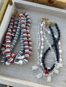 Two groups of beaded necklaces in a display tray; one set has blue, white, and red flat beads, the other has clear, dark blue, and striped beads with gold clasps.