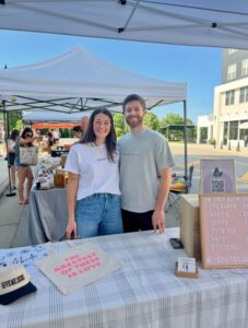Two people stand behind a market stall displaying shirts, tote bags, and other merchandise. A sign lists products and prices. Outdoor tents and other vendors are in the background.