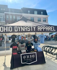 Three people stand behind a display of hats and shirts under a tent labeled "Ohio Dynasty Apparel" at an outdoor event in front of a building.