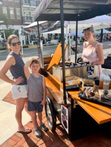 A woman and a boy stand next to a food cart vendor outdoors on a sunny day, with buildings and tents visible in the background.