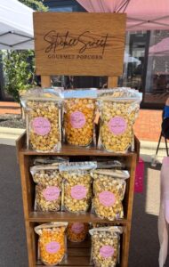 A display stand with several bags of gourmet popcorn in various flavors, labeled “Stickle Sweet Gourmet Popcorn,” set up at an outdoor market.