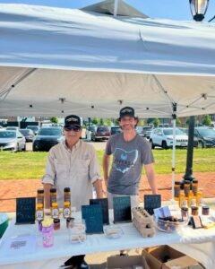 Two vendors stand behind a farmers market stall displaying eggs, baklava, creamed honey, hot honey, and jars of honey under a white canopy.