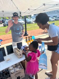 A man waves from behind a honey products booth at an outdoor market while a woman and a young girl in a pink dress sample honey.