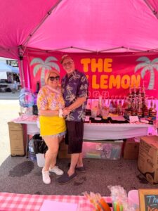 Two people stand smiling under a pink tent at a drink stand called "The Lemon," with flavored syrup bottles and supplies visible behind them.