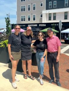 Four people stand smiling together on a sunny sidewalk in front of Sexton's Pizza, with apartment buildings and outdoor tents in the background.