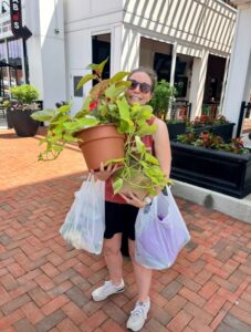 A woman stands outdoors on a brick walkway, holding a large potted plant and two plastic grocery bags with shops visible in the background.