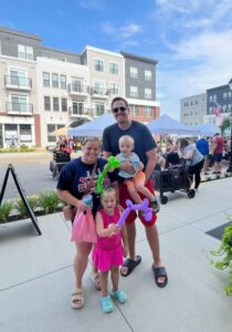 A smiling family of four with two young children and balloon animals pose on a sidewalk at an outdoor event, with tents and apartment buildings in the background.