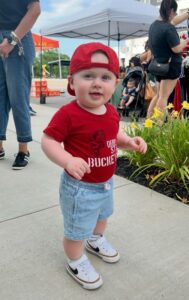 A toddler wearing a red cap, red t-shirt, and denim shorts stands on a sidewalk near yellow flowers at an outdoor event.