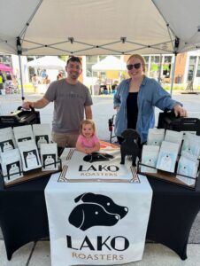 Two adults and a child stand behind a LAKO Roasters booth at an outdoor market, with coffee bags displayed on the table.