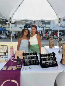 Two women stand behind a jewelry booth at an outdoor market, displaying bracelets, earrings, and rings on tables under a white canopy.