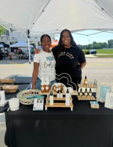 Two women stand behind a table displaying skincare products at an outdoor market under a white canopy tent.