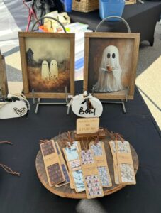 A display table featuring framed ghost artwork, a "BOO!" sign, and a wooden plate with bookmarks for sale at a market or fair.