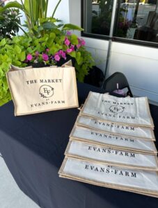 A display table with six canvas tote bags labeled "The Market at Evans Farm" arranged next to a plant.