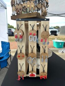 A wooden display stand holds various pairs of colorful handmade earrings on round brown cards, set up outdoors at a market.