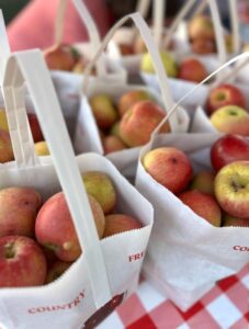 Several white paper bags filled with red and yellow apples are arranged on a red and white checkered tablecloth.