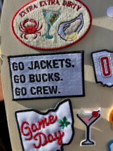 Various embroidered patches, including one with a crab, martini, and oyster, and another reading "GO JACKETS. GO BUCKS. GO CREW." are displayed on a board.