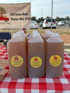 Plastic bottles of fresh cider from Country Road Farms are arranged on a red and white checkered tablecloth at an outdoor market.