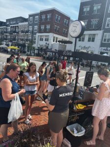 A group of people stand in line at an outdoor cart labeled "Little Farm" in a plaza with multi-story buildings in the background.