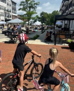 Two children on bikes pause on a brick walkway, observing a street market with tents, vendors, and people on a sunny day.