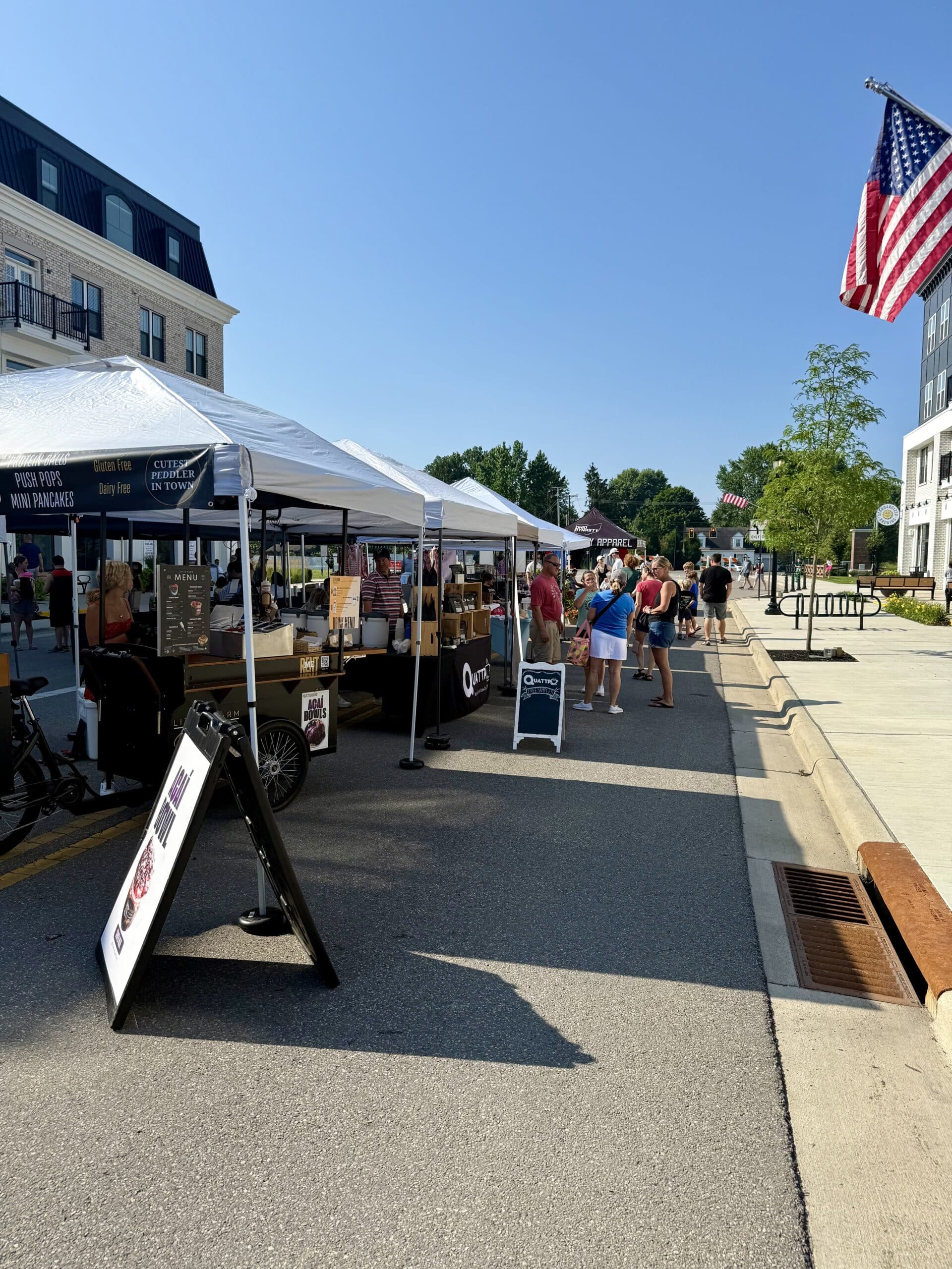 People browse vendor tents at an outdoor market on a sunny day, with an American flag on display and buildings lining the street.