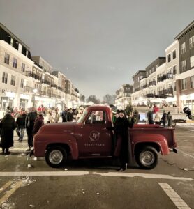 A red vintage Evans Farm pickup truck is parked on a city street at night, surrounded by people and modern buildings.