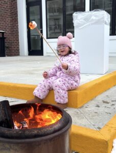 A young child in a pink snowsuit and hat sits on a curb roasting a marshmallow over a fire pit on a city sidewalk.