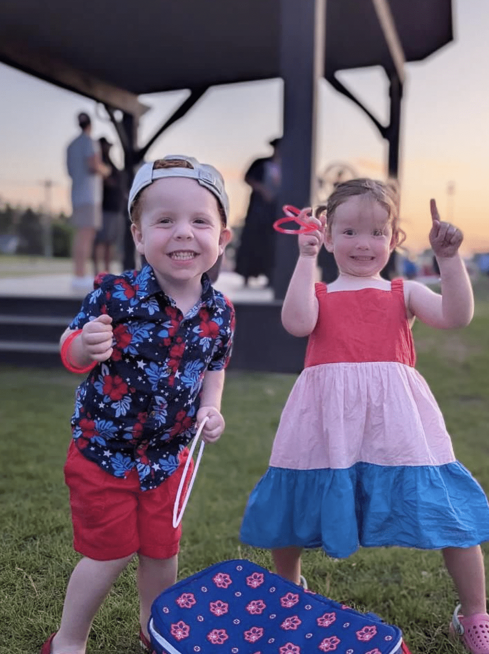 Two smiling young children stand on grass with a bag in front of them, holding glow sticks at an outdoor event near sunset.